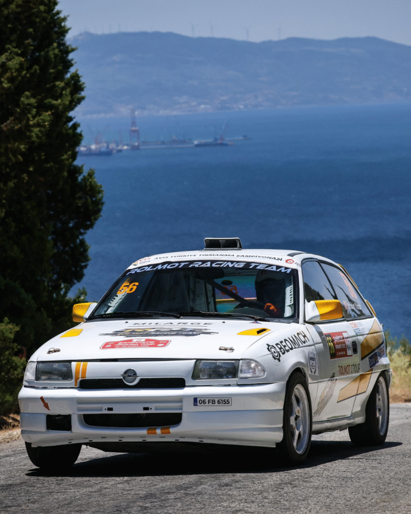 Rolmot Racing Team Opel Astra rally car climbing a coastal mountain road with the Aegean Sea in the background